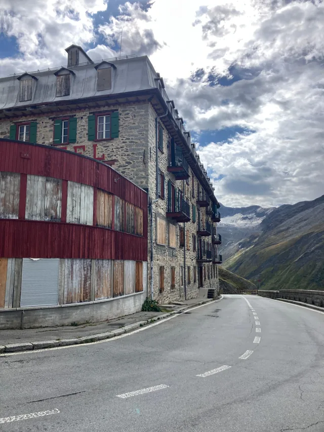 The weathered stone Hotel Belvedere with green shutters and a faded red wooden annexe beside the empty Furka pass road, the Rhône glacier visible in the mountainside behind
