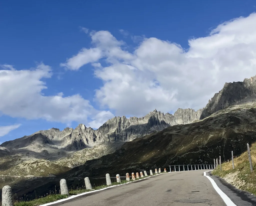 Empty Furka pass road curving along a mountainside lined with white and orange marker posts, beneath jagged granite peaks under a blue sky with white clouds