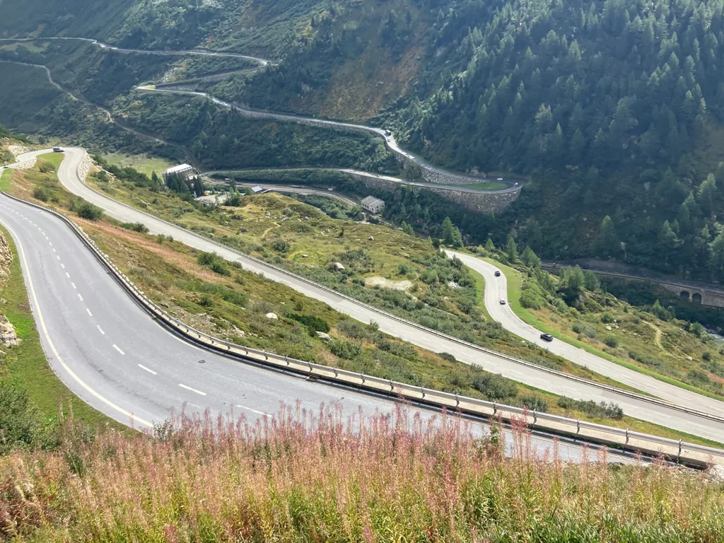 Sweeping hairpin roads climbing the Grimsel pass above Gletsch, with stone-walled switchbacks, a stone arch bridge and forested slopes, and the Furka pass road winding down the valley behind