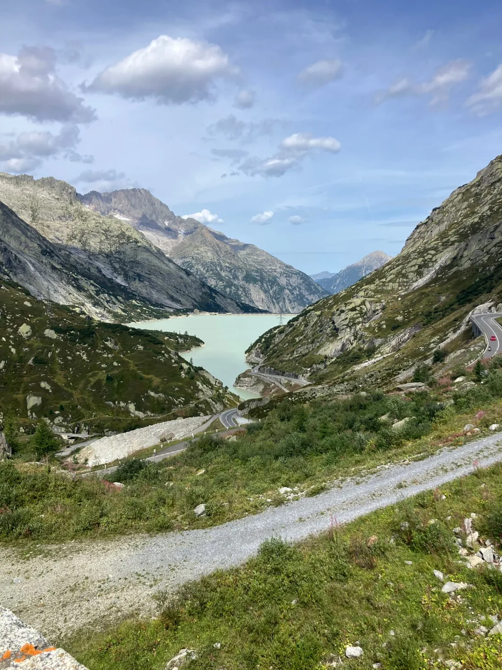 The turquoise Räterichsbodensee reservoir set between grey granite mountains, with the Grimsel pass road climbing the hillside on the right and a gravel track in the foreground