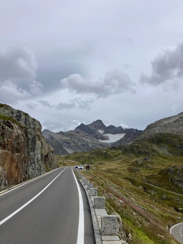 The Susten pass road climbing past stone marker posts over a rocky ridge, with a glacier-capped peak and a patch of blue glacier in the distance under grey clouds